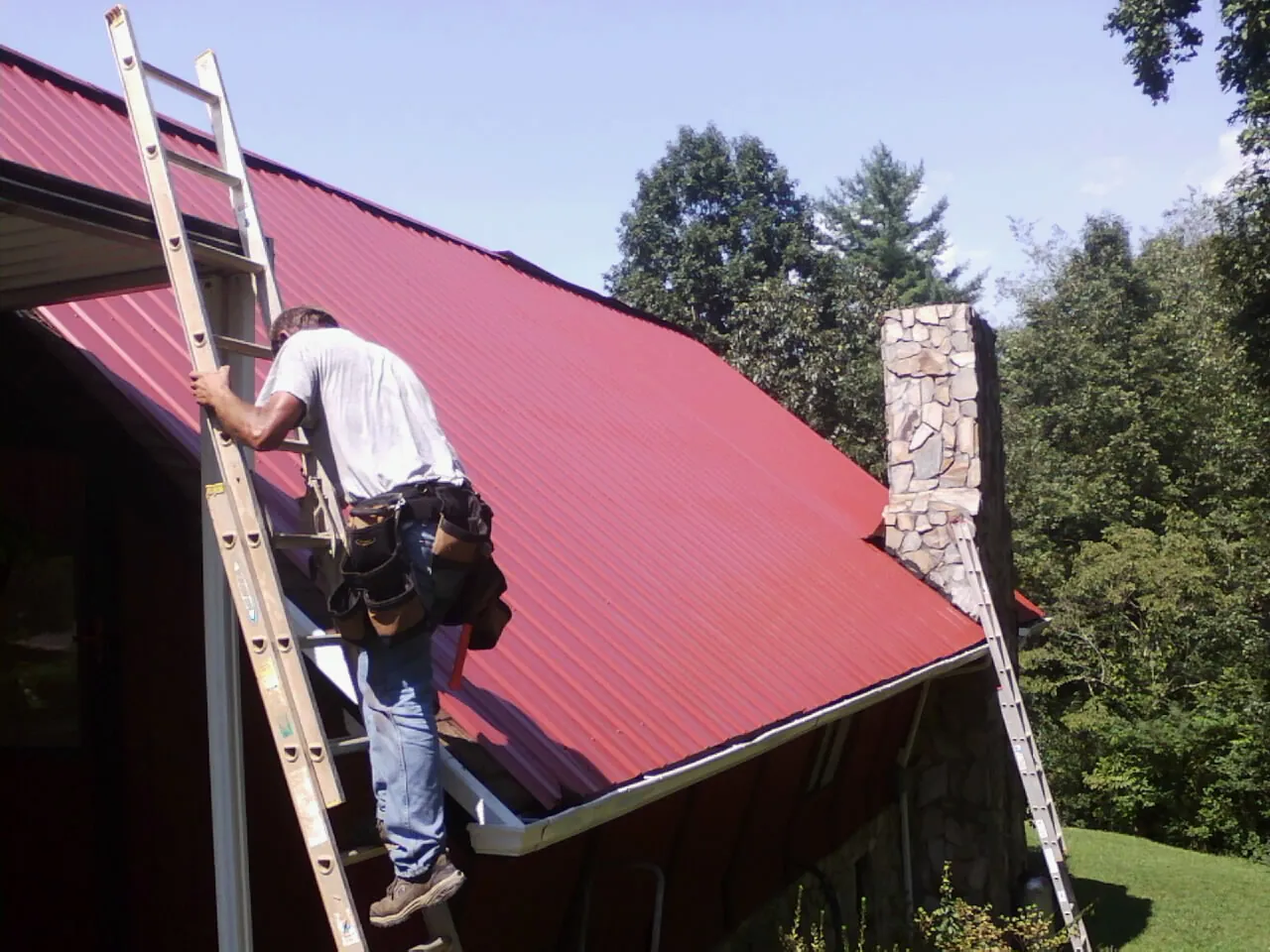 Roofer on a ladder beside a red metal roof and stone chimney — Soffit & Fascia Repair by Roofing Brothers in Bonanza